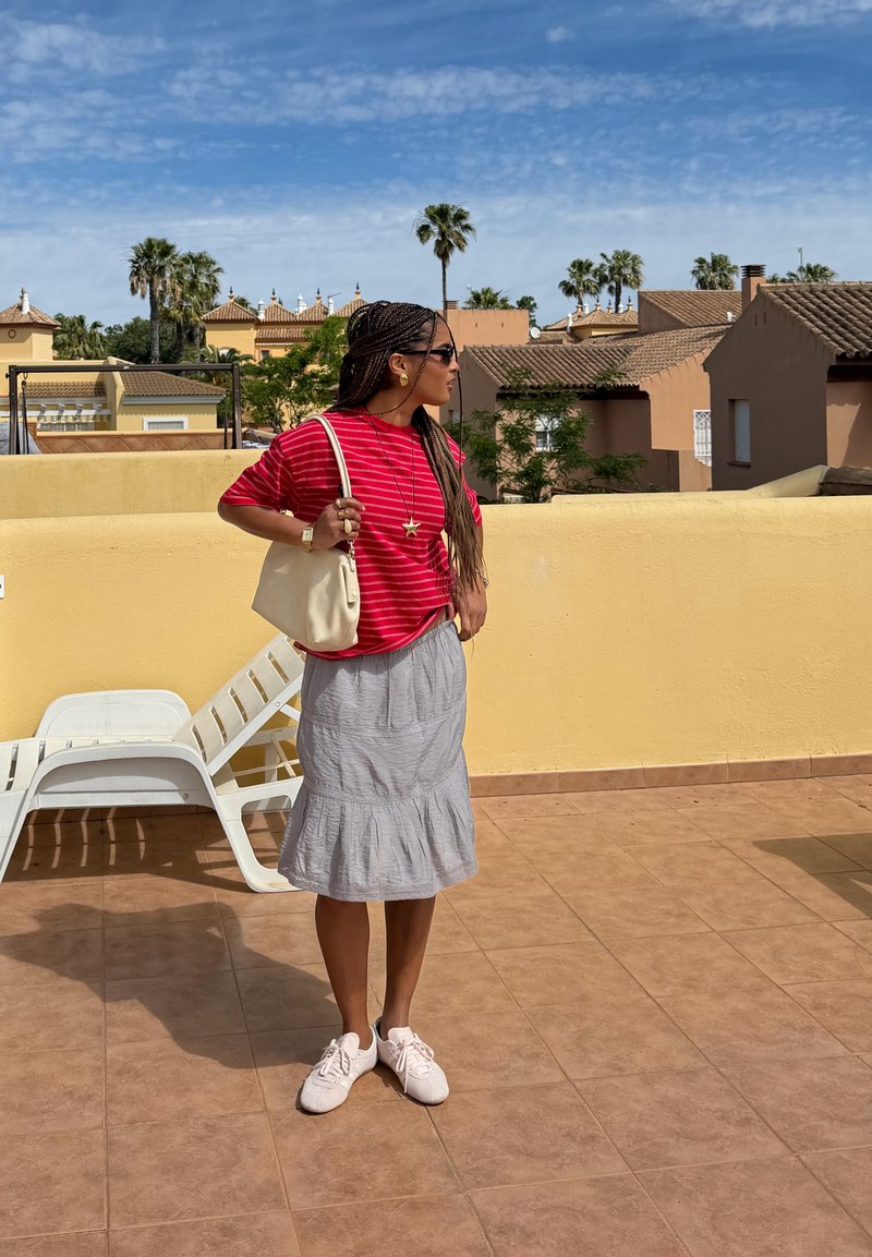 Femme aux cheveux tressés portant un haut rayé rouge, une jupe grise, des lunettes de soleil et des chaussures blanches, debout sur une terrasse ensoleillée avec des palmiers et des maisons en arrière-plan.