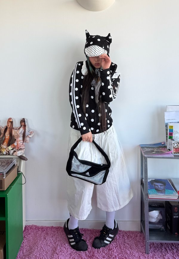 Persoană purtând un hanorac negru cu buline albe și urechi de animal, pantaloni albi largi, pantofi sport negri, ținând o geantă neagră Adidas.