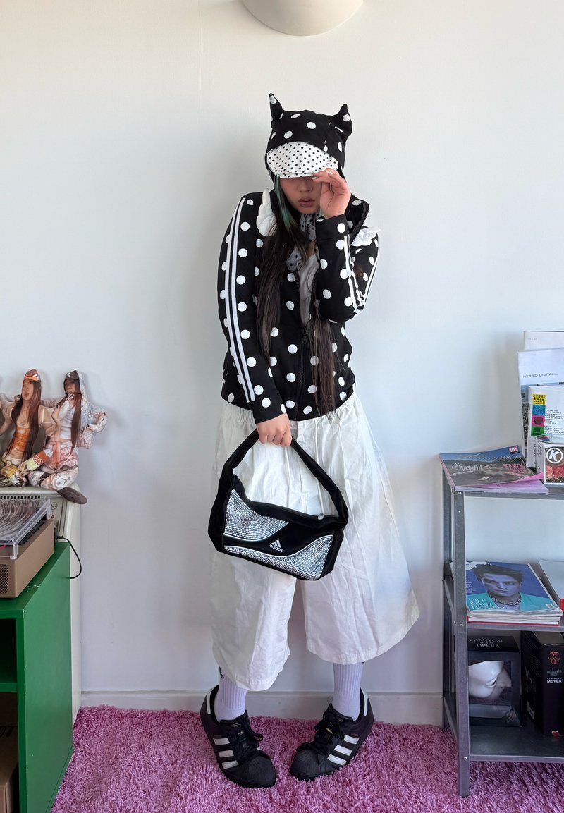 Persoană purtând un hanorac negru cu buline albe și urechi de animal, pantaloni albi largi, pantofi sport negri, ținând o geantă neagră Adidas.