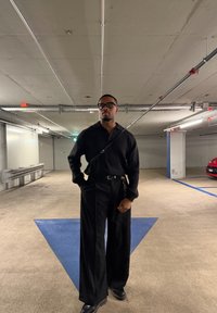 Čierny pletený sveter s dlhými rukávmi spojený so širokými čiernymi nohavicami. Doplnok zahŕňa opasok s kľúčenkou. Prostredie vnútorného parkoviska.