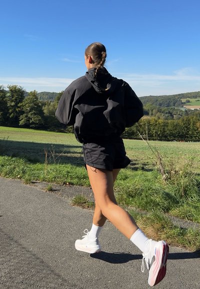 Sudadera negra de gran tamaño y pantalones cortos, combinados con zapatillas blancas y calcetines cortos, caminando por un sendero junto a campos de hierba bajo un cielo azul claro.