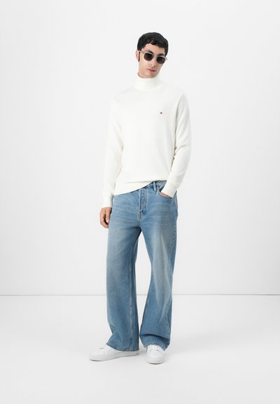 Suéter blanco de cuello alto con un pequeño logo, combinado con jeans de pierna ancha azul claro y zapatillas blancas. Modelo lleva gafas de sol.