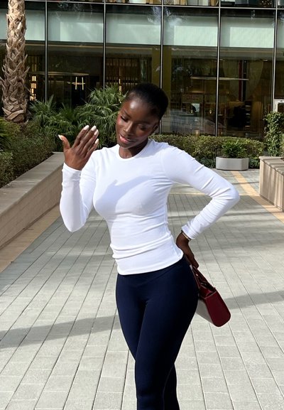 Mujer con blusa blanca de manga larga y pantalones negros, sosteniendo un bolso rojo, posando con una mano detrás de la espalda y la otra cerca del rostro al aire libre.