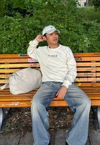Cremfarbener Sweatshirt mit Rundhalsausschnitt und "ellesse"-Logo, hellgraue weit geschnittene Jeans, weiße Kappe und beiger Reisetasche. Sitzt auf einer Holzbank.