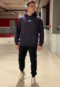 Marineblå hettegenser med logo foran, svarte cargobukser og beige joggesko med hvite detaljer. Glatt tekstur, avslappet passform og praktiske detaljer.