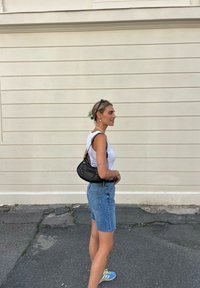 Bolsa preta com tachas decorativas, utilizada à tiracolo. Modelo com uma t-shirt branca sem mangas e calções de ganga, exibindo um vibrante acessório floral no cabelo.