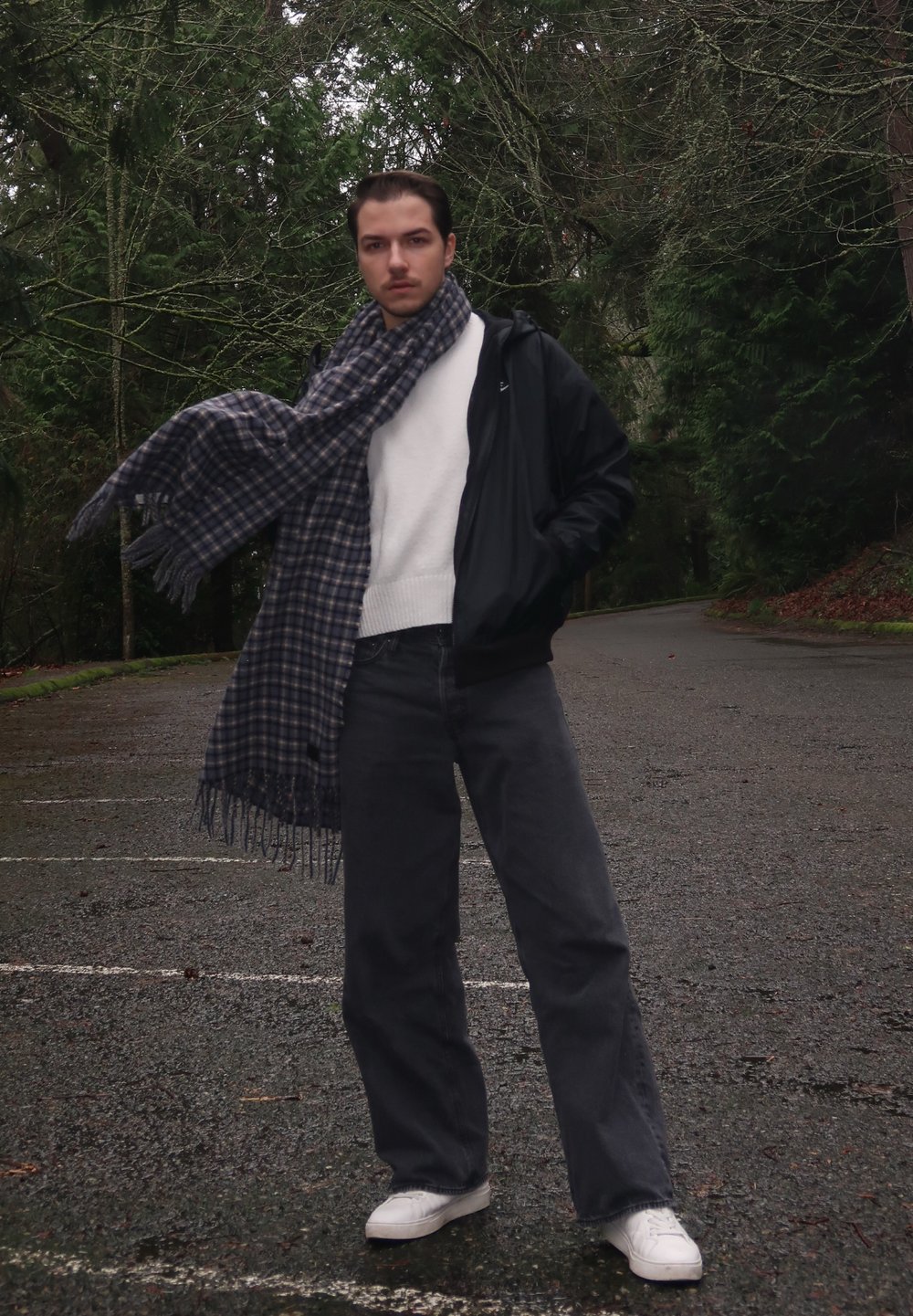 Une personne se tient dans une zone boisée, portant une veste noire zippée, un pull blanc, un pantalon gris foncé et des chaussures blanches. Une écharpe à carreaux bleue et grise est drapée autour de son cou.