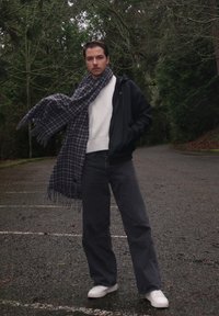 Une personne se tient dans une zone boisée, portant une veste noire zippée, un pull blanc, un pantalon gris foncé et des chaussures blanches. Une écharpe à carreaux bleue et grise est drapée autour de son cou.