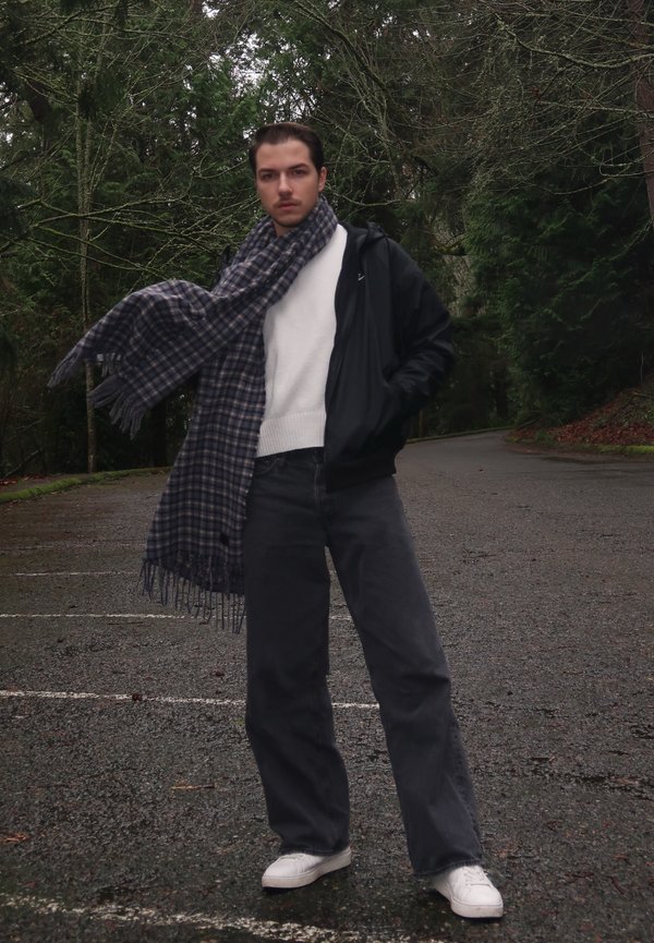 Egy személy áll egy erdős területen, fekete cipzáras kabátot, fehér pulóvert, sötétszürke nadrágot és fehér cipőt viselve. A nyakában kék és szürke kockás sál van drapírozva.