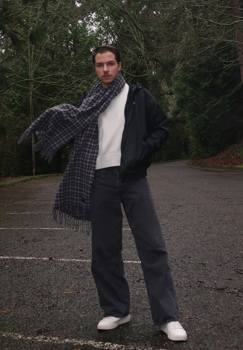 Een persoon staat in een bosrijk gebied, gekleed in een zwarte jas met ritssluiting, een witte trui, donkergrijze broek en witte schoenen. Rond de nek hangt een blauw-grijs geruite sjaal.