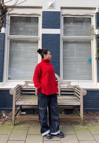 Maglione a collo alto rosso a coste con tasche laterali, abbinato a pantaloni in denim scuro con cuciture a contrasto e orli arrotolati, indossato con calzature nere.