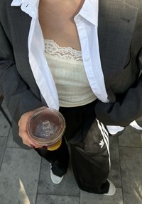 Blazer cinza sobre uma camisa branca e uma camisola leve com renda, combinada com calças de fato pretas com riscas brancas e o logótipo da Adidas. Copo transparente com tampa.