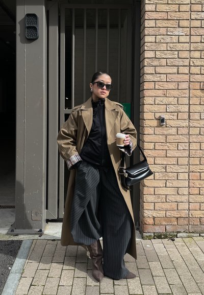 Mujer con abrigo largo beige y pantalones anchos negros con rayas sostiene una taza de café y un bolso negro, de pie contra una pared de ladrillos.