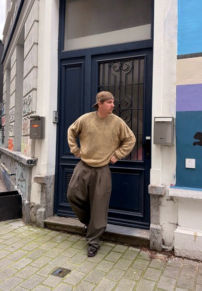 Joven con suéter beige, pantalones marrones holgados y gorra a cuadros, de pie con las manos en la cintura frente a una puerta oscura en una calle cubierta de grafitis.
