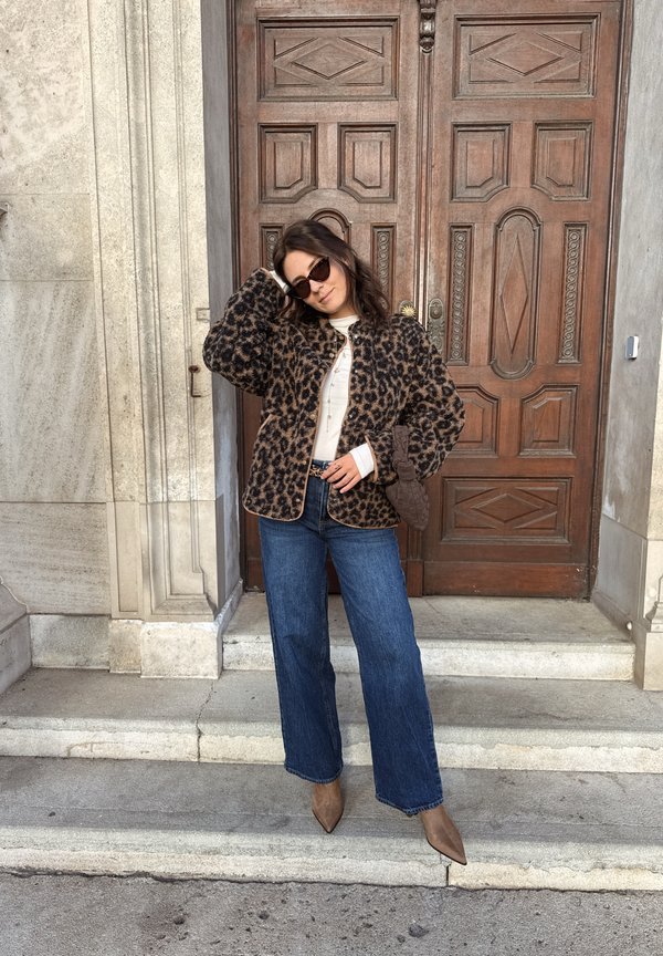 Mujer con chaqueta de estampado de leopardo, vaqueros azules, botas marrones puntiagudas y gafas de sol, de pie sobre escalones de piedra frente a puertas dobles de madera.