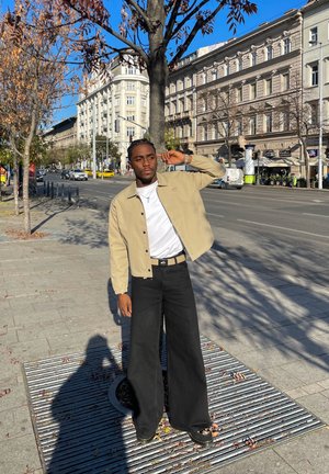 Chaqueta beige sobre una camisa blanca, combinada con pantalones de pierna ancha negros y zapatos negros. De pie sobre un pavimento texturizado con un árbol cercano.