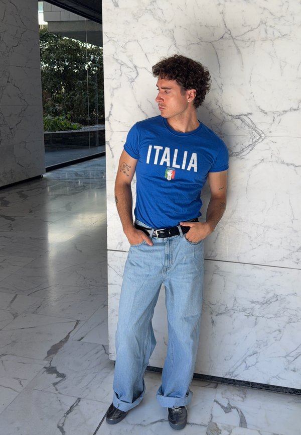 Uomo con capelli ricci che indossa una maglietta blu "ITALIA" e jeans azzurri, in piedi contro un muro di marmo bianco con le mani in tasca.