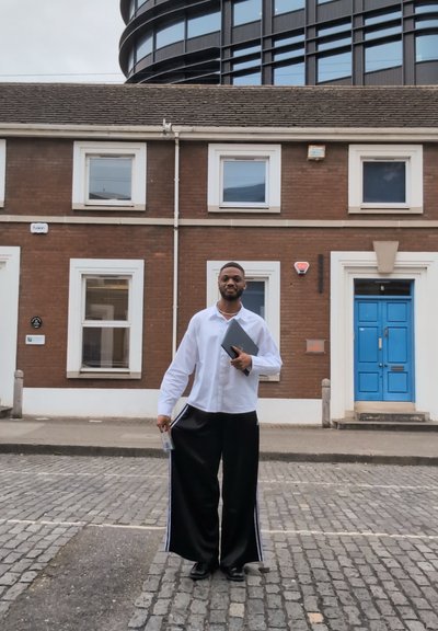 Una persona se encuentra frente a un edificio de ladrillo marrón, vistiendo una camisa blanca y pantalones anchos negros con rayas blancas. Sostiene una tableta y una identificación.