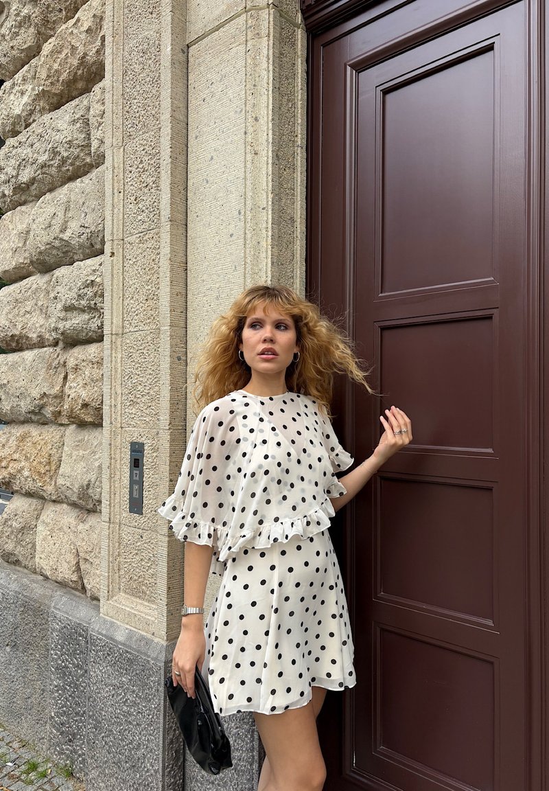 Valkoinen pilkullinen mekko, jossa on röyhelöhelma ja -hihat, yhdistettynä mustaan laukkuun. Taustalla on tekstuuri-kiviseinä ja ruskea ovi.