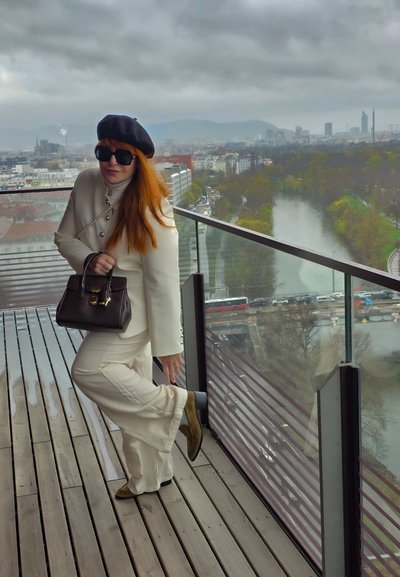 Mujer con traje blanco y boina negra ajusta zapato en balcón con vistas al río, la ciudad y el cielo nublado.