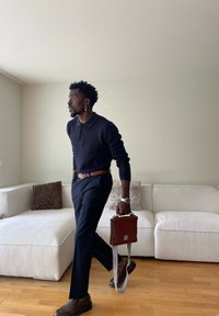 Mann in dunklem Pullover und Hose geht drinnen, trägt eine braune Ledertasche mit gestreiftem Riemen, nahe einem weißen Sofa mit gemusterten Kissen.