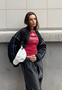 Jeune femme appuyée contre un mur, portant un haut rouge court, une veste noire, un pantalon noir ample, avec une casquette blanche suspendue à son épaule.