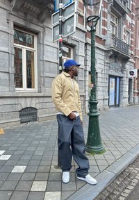 Beige zip-up jack over een wit shirt, losse blauwe denim broek, en witte sneakers met blauwe accenten, staande bij een groene straatlantaarn.