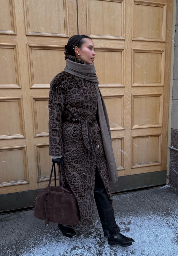 Frau mit Leopardenmuster-Mantel und Schal geht auf schneebedecktem Gehweg und trägt eine dunkle Handtasche, vor einem Hintergrund aus holzvertäfelter Tür.