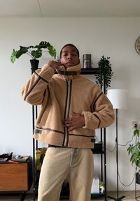 Veste polaire beige avec un col montant, des fermetures éclair et des accents marron contrastants, présentant une coupe décontractée et une surface texturée.