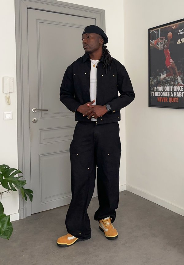 Uomo con dreadlocks che indossa giacca e pantaloni neri, scarpe da ginnastica gialle e bianche, in piedi all'interno vicino a una porta grigia e a un poster motivazionale di basket.