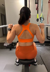 Femme en tenue de sport orange réalisant un exercice de tirage latéral assis dans une salle de sport avec des bandes de résistance suspendues à proximité.
