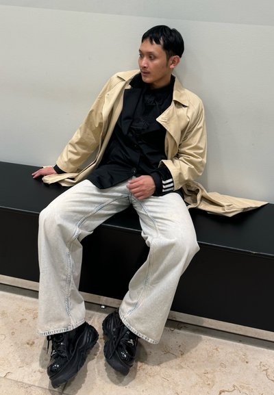 Abrigo trench beige sobre una camiseta negra, pantalones de mezclilla anchos de color claro y zapatillas negras gruesas. Sentado en un banco negro contra una pared clara.