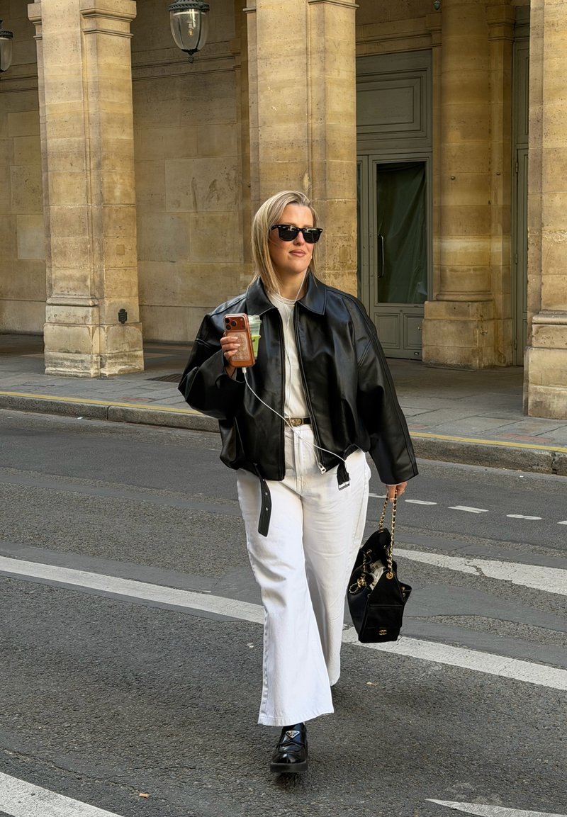 Femme portant une veste en cuir noire et un pantalon blanc marche dans la rue en tenant une boisson, un téléphone et un sac à main noir avec une chaîne dorée.