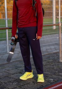 Atlet bærer en rød langærmet trøje og marineblå bukser, og holder en ski. Skoene er lysegule med grå detaljer. Udendørs baggrund med hegn.