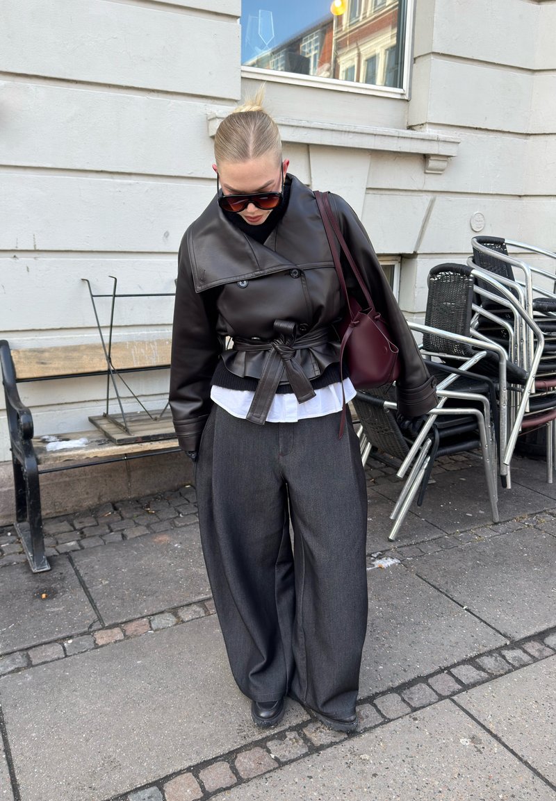 Femme portant une veste en cuir noire ceinturée, un pantalon large gris, de grandes lunettes de soleil et un sac bordeaux, debout sur le trottoir près de chaises empilées.
