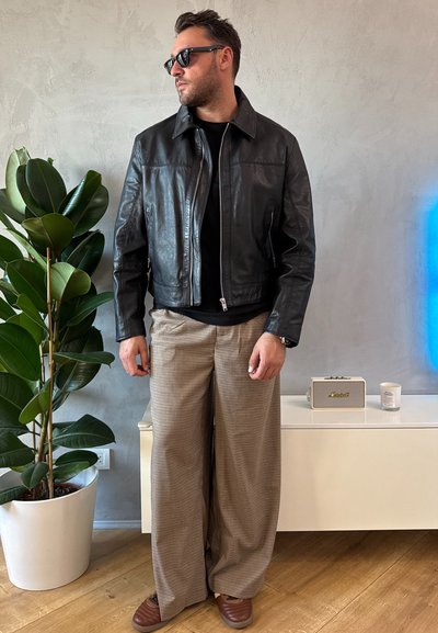 Chaqueta de cuero negra, camisa negra, pantalones anchos de cuadros beige, zapatillas marrones, gafas de sol; ambiente interior con una planta y objetos decorativos.