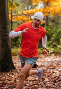 Juoksija aktiivivaatteissa, päällään kirkkaanoranssi lyhythihainen paita, harmaat shortsit ja valkoinen pipo, juoksee metsäisessä ympäristössä, jonka maata peittää syksyn lehdet.