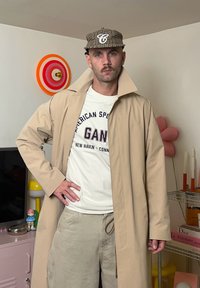 Casaco trench bege, t-shirt branca com o texto “GANG”, calças verde-oliva, boné com padrão. O fundo inclui decoração e uma televisão.