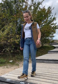 Chaleco de forro polar marrón con detalles en verde, camiseta blanca con logo, jeans azul claro y botas de montaña negras con detalles amarillos, caminando por un sendero de madera.