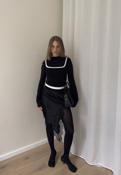 Mujer de pie en interiores con un vestido negro con ribete blanco, medias y zapatos negros, sosteniendo un bolso negro junto a una pared y cortina claras.