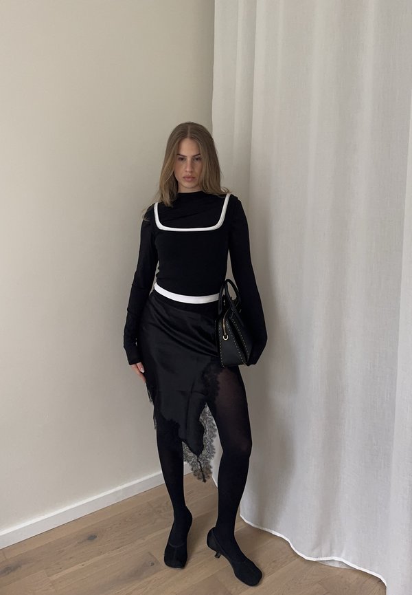 Femeie stând în interior purtând o rochie neagră cu garnitură albă, ciorapi negri și pantofi, ținând o geantă neagră în fața unui perete și a unei draperii deschise la culoare.