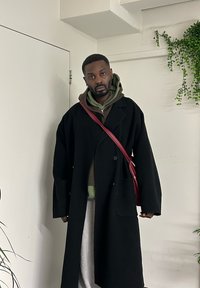 Homme portant un long manteau noir par-dessus des sweats à capuche superposés, avec un sac bandoulière rouge, debout à l'intérieur près d'une porte blanche et de plantes vertes.