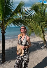 Combinaison-pantalon à rayures noires et blanches avec un col halter, associée à un chapeau de paille à larges bords, sur un fond de palmiers et de paysage de plage.