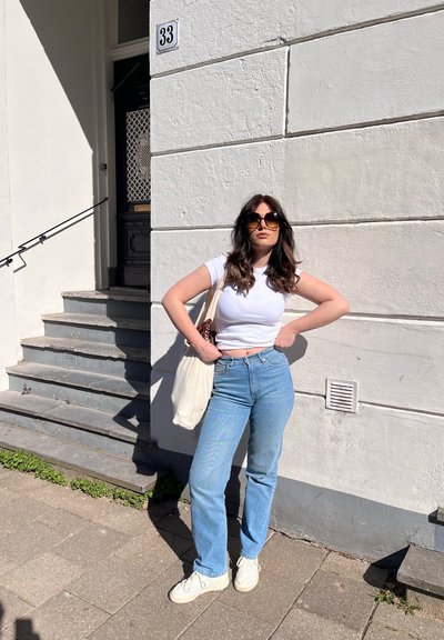 Mujer con gafas de sol, camiseta blanca, vaqueros azules y deportivas blancas, de pie con las manos en las caderas junto a un edificio blanco con el número 33 a plena luz del día.