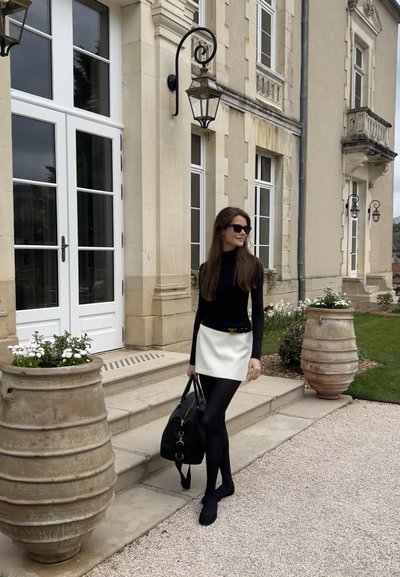 Blusa negra de manga larga, falda mini blanca, medias negras y zapatos planos, llevando un gran bolso negro. Escalera de piedra y flores en macetas al fondo.