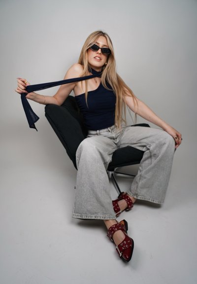 Top negro de malla, pantalones vaqueros anchos gris claro y zapatos de tacón rojos con herrajes plateados. Sentada en una silla moderna negra con un fondo neutral.