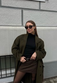 Femme portant des lunettes de soleil foncées, un manteau vert olive, un haut noir, une jupe marron, debout contre un mur gris clair texturé, les mains sur sa ceinture.