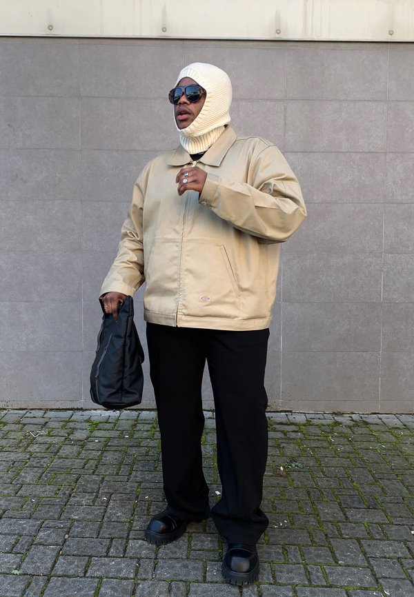 Beige oversized jacket, black trousers, chunky black shoes, white knitted balaclava, holding a black bag, sunglasses reflecting the surroundings.