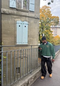 Grå stenbyggnad med blå fönsterluckor och runda metalldetaljer. En person i grön tröja och svarta byxor går längs ett grått räcke.