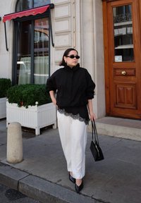 Musta ylisuuri takki, valkoiset housut pitsireunalla, mustat korot ja aurinkolasit, pitäen mustaa käsilaukkua. Kaupunkimaisema, jossa on koristeellisia ruukkuja.